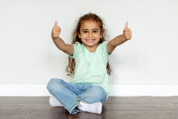 Portrait of pretty smiling child girl showing thumbs up at camera while sitting on floor over light wall background, kid gesturing sign of approval