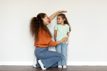 Young european mother measuring height of her daughter near white wall at home, full length shot