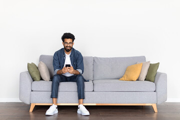 Casual Indian Male Enjoying Time on Couch with Phone - obrazy, fototapety, plakaty