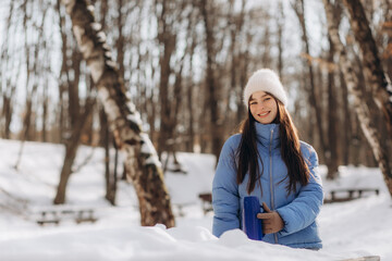 Portrait of young woman in warm clothes in winter forest. Drinking hot tea outdoors from thermos. Hiking.