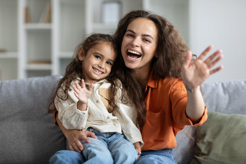 Excited european woman hugging her daughter and waving hands at camera, sitting on sofa in living...