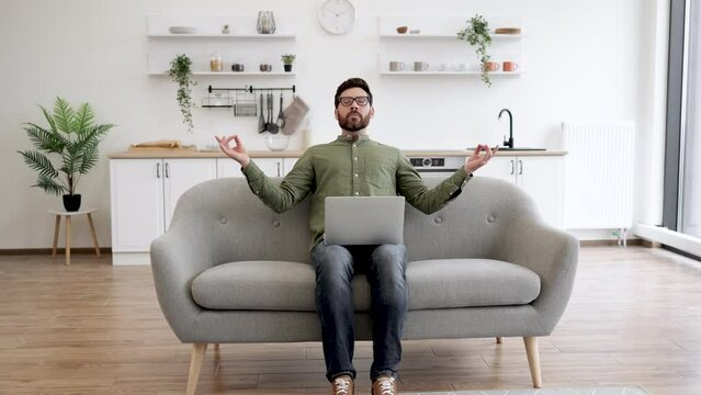 Peaceful caucasian man in glasses and casual attire sitting on couch with wireless laptop on knees and meditating. Bearded young guy with fingers in mudra gesture relieve stress and regaining energy.