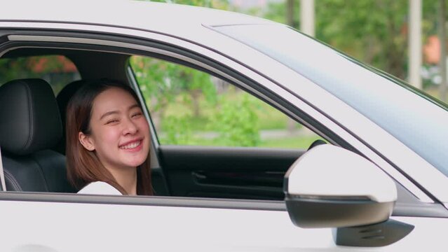 Happy Asian woman sitting in car behind steering and wave hand. Concept of happy driver, car insurance service. Customers excited after buying new car, new drivers excited for successful car license