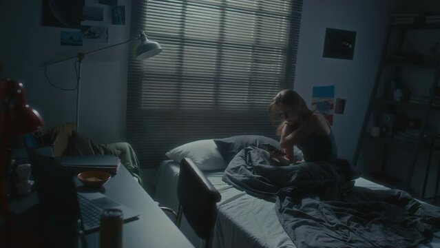 High angle shot of young woman sitting alone in her bedroom during sleepless night, using smartphone and feeling sad because of cyberbullying or doomscrolling