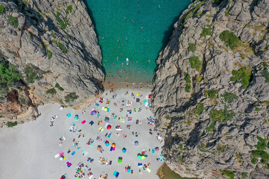 Aerial view of Sa Calobra beach with people swimming in the clear blue water and laying on the rocky sand with towels and umbrellas between two mountains in Balearic Islands, Spain.