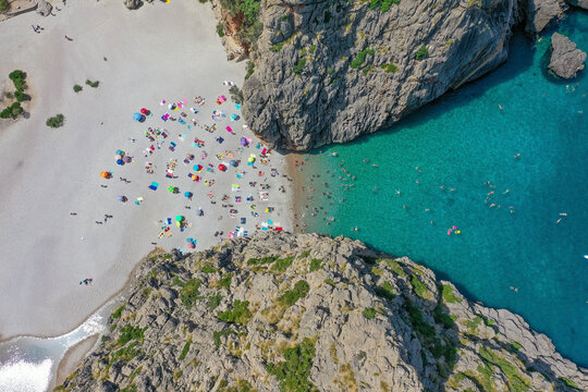 Aerial view of Sa Calobra beach with people swimming in the clear blue water and laying on the rocky sand with towels and umbrellas between two mountains in Balearic Islands, Spain.