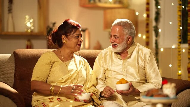 Indian aged Husband feeding sweets to her wife on the diwali festival - Colorful background with lights.wife holding sweets.they both are sitting on the sofa.