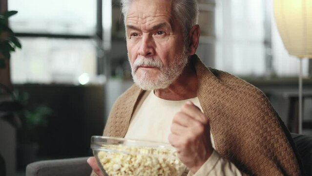 Overjoyed gray haired senior man watch sport game and comments emotionally at cozy home Handsome mature male fan with bowl of popcorn enjoying the weekend and cheers for favourite club alone indoors