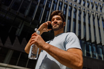 Fototapeta na wymiar Smiling man tourist drink water and talking by phone while standing on city skyscrapers background
