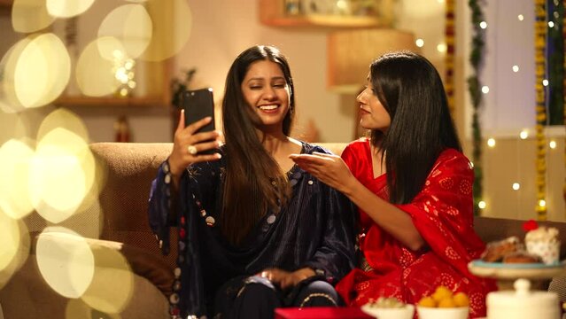 Two indian sister happily waving and chatting on a video call during Diwali festival.they both are sitting on the sofa. Diwali decorations,wearing traditional dress.