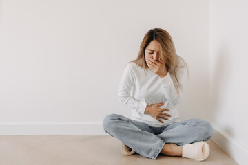 Fototapeta na wymiar Asian Thai woman feeling sick and queasy, nausea and vomiting, grabbing stomachache cramp and covering mouth, wear white sweater, sitting on floor in white apartment room in winter.