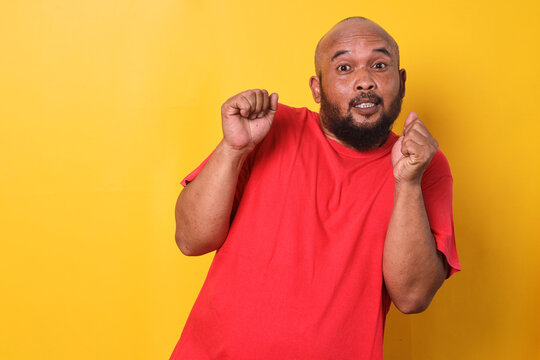 Portrait of scared bald man in red t-shirt clenching fist feeling afraid over yellow background