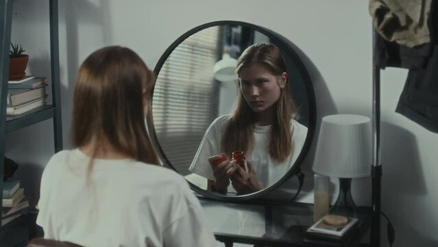 Sad girl with suicidal thoughts holding pills and looking in mirror during day at home