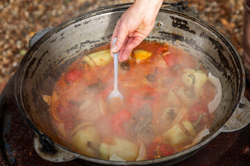 A woman cooks in a cauldron over an open fire and stirs with a spoon. Picnic in nature. Close-up.