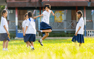 Fototapeta premium Close up group of Asian girls wear student uniform enjoy to play with jump across rubber string in grass field area of school.