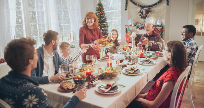 Diverse Group of Relatives and Friends Sitting Together Behind a Dining Table with Tasty Meals and Festive Decorations. Senior Woman Bringing a Grilled Chicken, Creating a Joyful Holiday Atmosphere