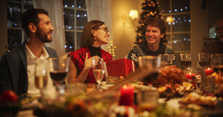 Handsome Boyfriend Presenting a Festive Gift to His Beautiful Surprised Girlfriend. Family and Friends Gather at Home in the Evening for a Traditional Christmas Dinner with a Turkey Roast Feast
