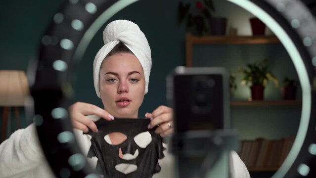 Young woman in towel records video tutorial at home applies sheet moisturizing mask on her face shoots broadcast live video to social network. Blogging, skin care and beauty
