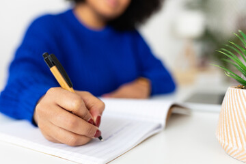 Close-up view of young adult woman taking notes in notebook while using laptop at home. Focus on hand. Business and education concept.