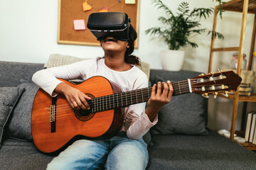 Obraz premium African american teenage girl sitting on couch in her room and learning to play guitar using VR glasses to immerse herself in metaverse