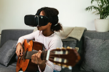 African american teenage girl sitting on couch in her room and learning to play guitar using VR...