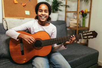 African american teenage girl sitting on couch in her room and learning to play guitar