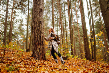 Beautiful young tourist with a backpack is having fun in the autumn forest, throwing fallen leaves. Woman walks along a colorful path in the forest. Freedom, travel.