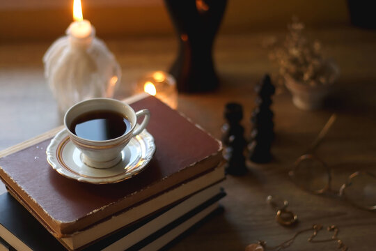 Vintage books, reading glasses, gold pen, hand model, gold jewelry, scented candles, flowers, chess pieces and cup of tea or coffee on the table. Dark academia concept. Selective focus.
