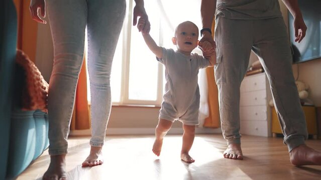 baby with parents first steps. happy family kid dream concept. baby son takes first steps holds family hands silhouette against window indoors. cute baby toddler learning walk with lifestyle family