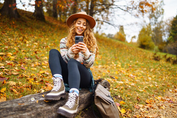 Young curly woman in a hat with a phone in an autumn park among yellow fallen leaves. Smiling woman using smartphone outdoors. Concept for vacation, weekend, technology.