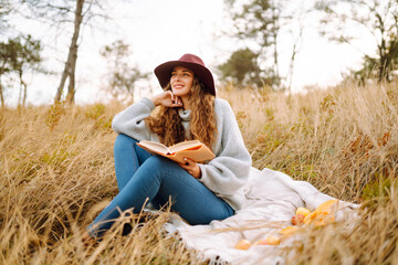 Naklejka na ściany i meble Young woman in a cozy sweater and hat enjoys nature, reading a book while sitting on a blanket in an autumn park. A beautiful woman is having a picnic outdoors.