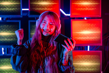 Winning. Confused Asian woman wearing gaming headphones feeling excited at gaming room, Happy Gamer playing video game online with smart mobile phone with neon lights raises hands to wins celebrating