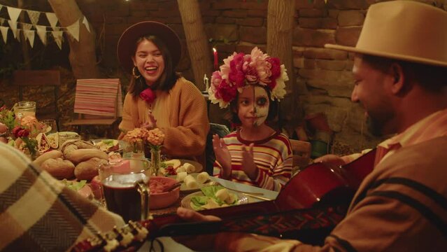 Waist up of delighted Hispanic man in camel hat and poncho playing guitar to family while sitting together at festively set dinner table celebrating Day of the Dead in cozy backyard at night