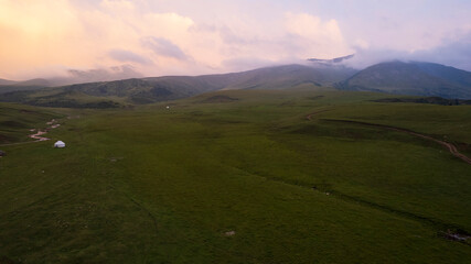 Pink-purple sunset in the mountains with green fields. There is a white yurt, a mountain river runs. Huge clouds. A herd of horses is walking. An SUV next to the mud and a tent. Tourists gather