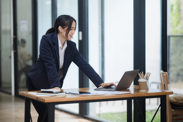 Asian woman working on laptop and standing at office desk.