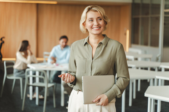 Smiling middle aged businesswoman holding laptop working in modern office, standing, looking away