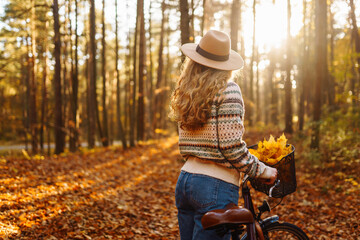 A beautiful woman in a hat and sweater rides a bicycle in an autumn park. Active tourist enjoying...