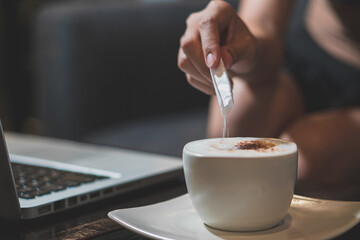 Closeup image of a hand holding a cup of hot latte coffee with laptop on wooden table in cafe