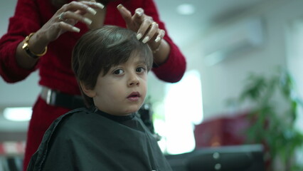 Little boy sitting at hair salon chair looking at his own mirror reflection wearing black cape getting his haircut, professional hairstylist drying hair