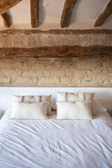 interior of rustic rural room, bed with white sheets and cushions, front view of headboard with white shelf and rustic stones of European rural house.