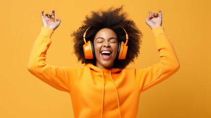 Happy young Afro American woman carried away with music dances carefree with arms raised keeps eyes closed wears stereo headphones on ears dressed in orange jumper isolated over white background