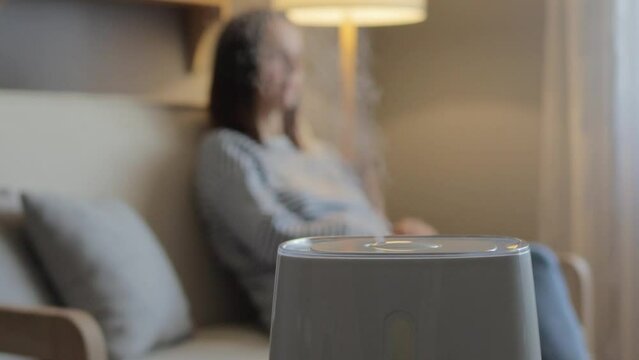 Modern air humidifier during relax or rest, blurred young woman enjoying aromatherapy steam scent from essential oil diffuser comfortable in living conditions room.
