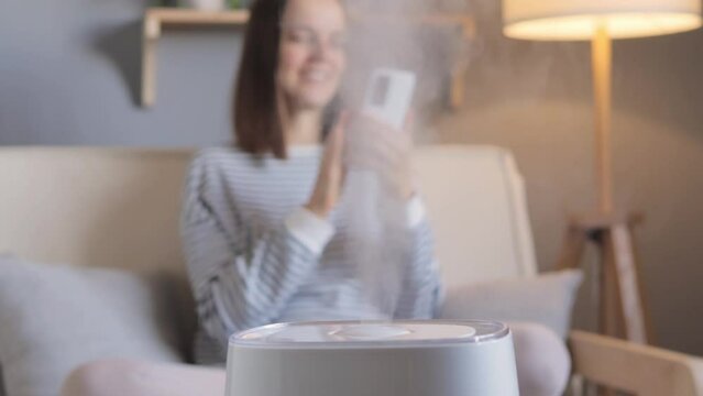 Pretty smiling happy woman sitting near air purifier and moisturizer appliance on couch enjoying air quality in her apartment using smartphone chatting online.