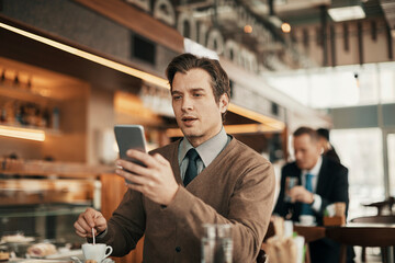 Young businessman using a smartphone while sitting in a cafe and stirring a cup of coffee