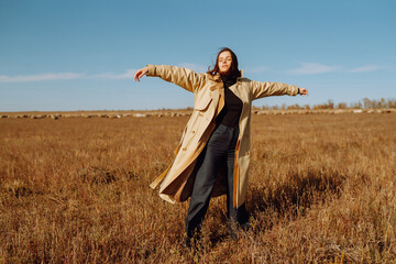 Young woman in a coat standing in a field among dry grass, enjoying nature. Urban style and street fashion. Concept of nature, fashion, freedom. Lifestyle.