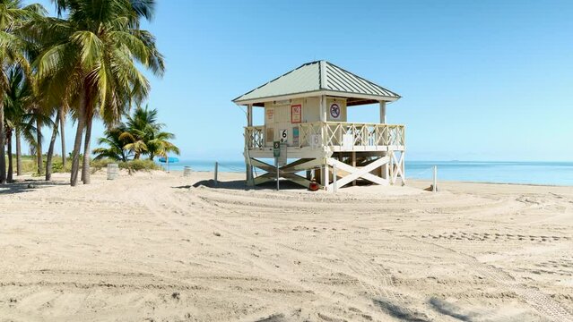 push in footage of a white wooden lifeguard tower on a sandy white beach with lush green palm trees and blue ocean water at Crandon Park in Key Biscayne Florida USA