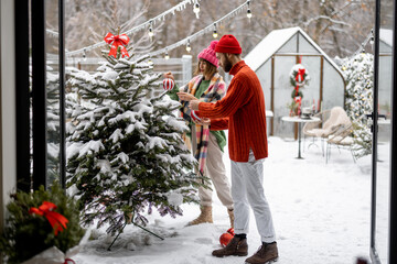 Man and woman decorate Christmas tree with festive balls, while preparing for a winter holidays at snowy backyard of their house. Happy family celebrating New Year's holidays