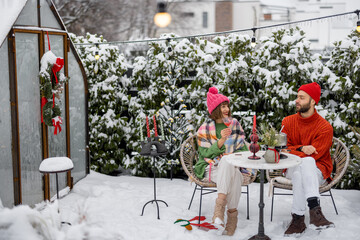 Man and woman have romantic dinner, while sitting together by the table at beautifully decorated snowy backyard. Young family celebrating winter holidays