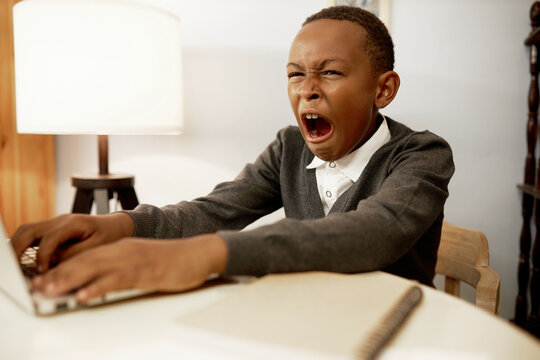 Indoor portrait of tired exhausted sleepy african american school child using laptop sitting at table in his room, studying online in morning, yawning typing on keyboard, doing exercises