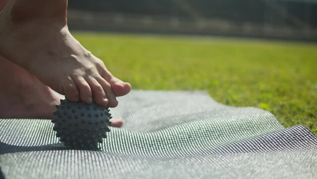 Young woman doing Myofascial Release for feet using a little ball on the sport mat on green grass close up. Massage for legs. Stretching, yoga outdoor